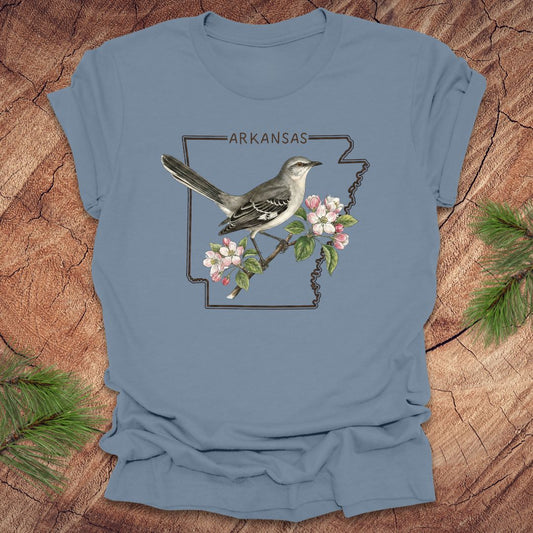 Blue t-shirt with a bird and floral design on an Arkansas map, placed on a wooden surface.