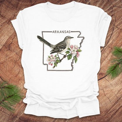 White t-shirt with Arkansas state design featuring a bird and flowers on a wooden surface.