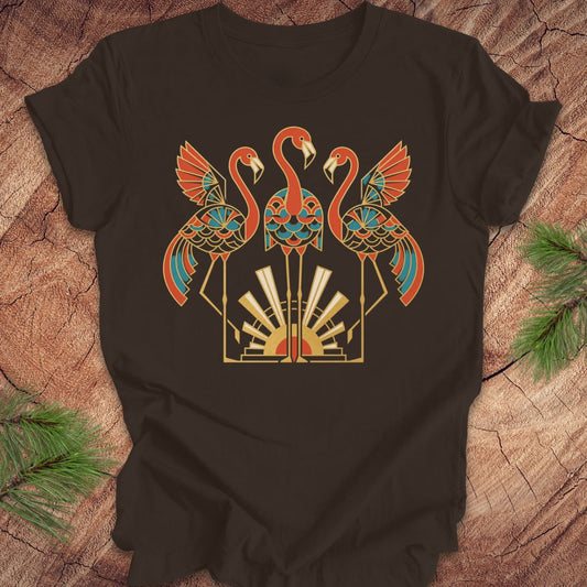 A brown short-sleeve t-shirt with a stylized graphic of three flamingos in orange, teal, and gold, laid flat on a rustic wood background with pine needles in the corners.