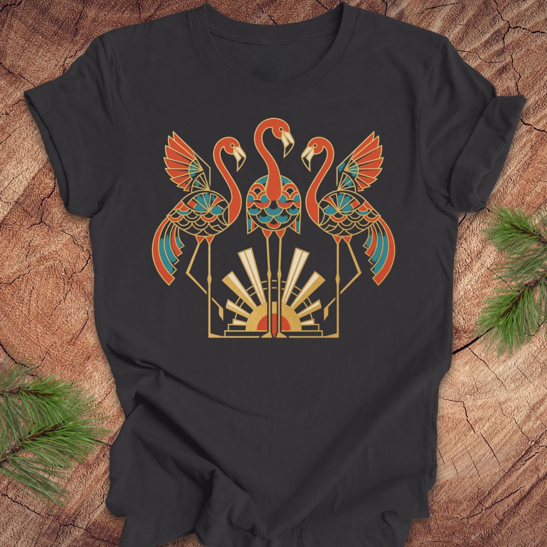 A dark gray short-sleeve t-shirt with a colorful graphic of three stylized flamingos in orange, teal, and gold, laid flat on a rustic wood background with pine needles in the corners.