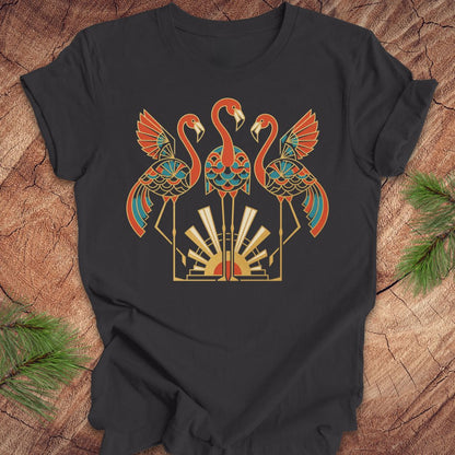 A dark gray short-sleeve t-shirt with a colorful graphic of three stylized flamingos in orange, teal, and gold, laid flat on a rustic wood background with pine needles in the corners.