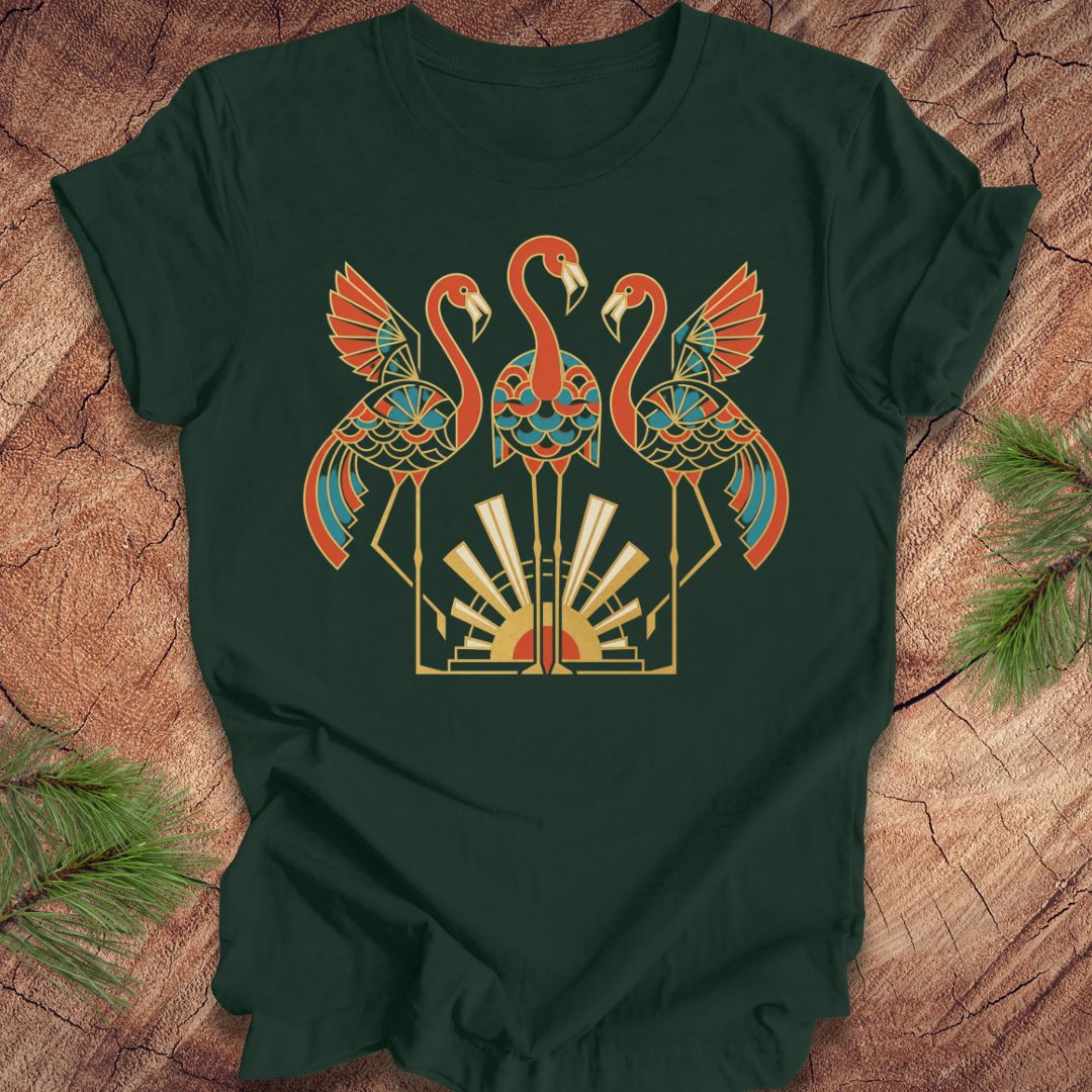 A dark green short-sleeve t-shirt laid flat on a rustic wood surface, featuring a large central graphic of two flamingos and a geometric sunburst design in orange, teal, cream, and gold tones. Pine branches are placed at the corners of the frame.