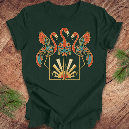 A dark green short-sleeve t-shirt laid flat on a rustic wood surface, featuring a large central graphic of two flamingos and a geometric sunburst design in orange, teal, cream, and gold tones. Pine branches are placed at the corners of the frame.