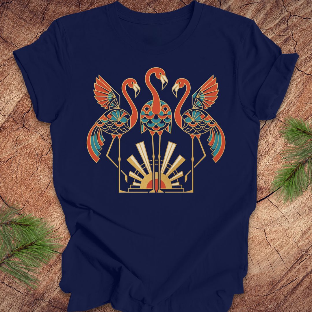 A navy blue short-sleeve t-shirt with a colorful graphic of three stylized flamingos in orange, teal, and gold, laid flat on a rustic wood background with pine needles in the corners.