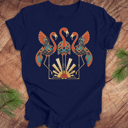 A navy blue short-sleeve t-shirt with a colorful graphic of three stylized flamingos in orange, teal, and gold, laid flat on a rustic wood background with pine needles in the corners.