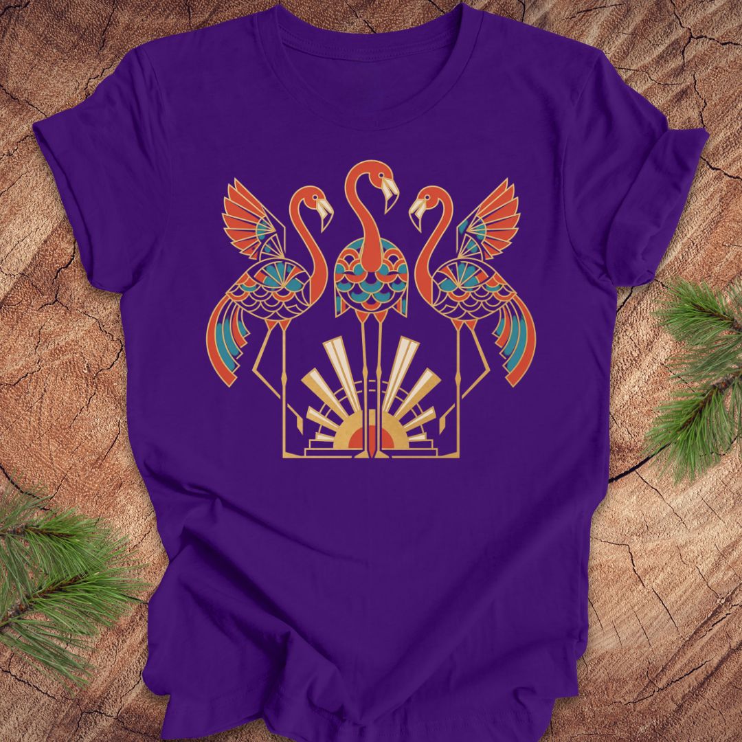 A purple short-sleeve t-shirt with a colorful graphic of three stylized flamingos in orange, teal, and gold, laid flat on a rustic wood background with pine needles in the corners.