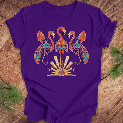 A purple short-sleeve t-shirt with a colorful graphic of three stylized flamingos in orange, teal, and gold, laid flat on a rustic wood background with pine needles in the corners.