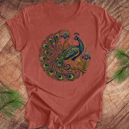 A terracotta-colored short-sleeve t-shirt featuring a detailed graphic of a peacock with a fanned tail and blue flowers, laid flat on a rustic wood background with pine branches.