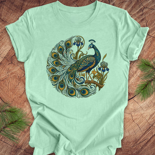 A mint green short-sleeve t-shirt with a detailed graphic of a peacock in a circular composition, laid flat on a rustic wood background with pine needles in the corners.