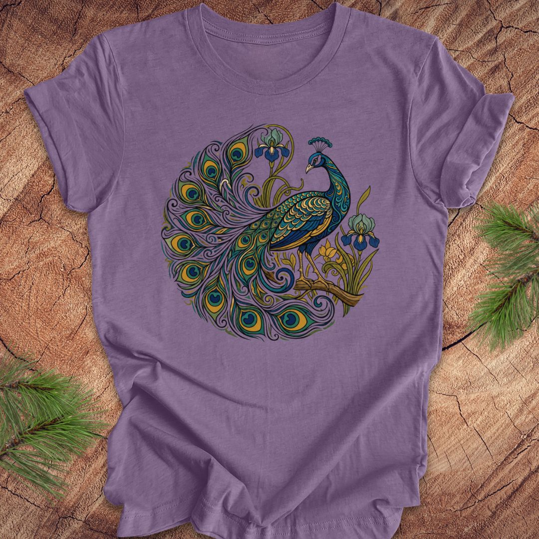 A lavender purple short-sleeve t-shirt featuring a detailed graphic of a peacock with a circular tail pattern in shades of teal, green, blue, and gold. The t-shirt is laid flat on a rustic wood slice background with pine needles visible in the corners.