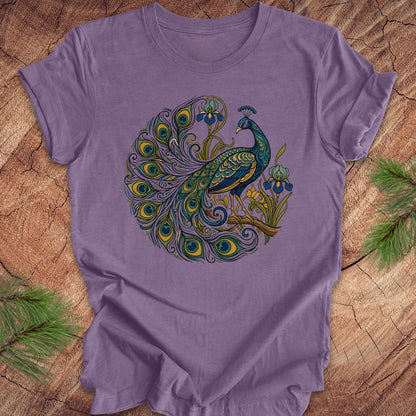 A lavender purple short-sleeve t-shirt featuring a detailed graphic of a peacock with a circular tail pattern in shades of teal, green, blue, and gold. The t-shirt is laid flat on a rustic wood slice background with pine needles visible in the corners.