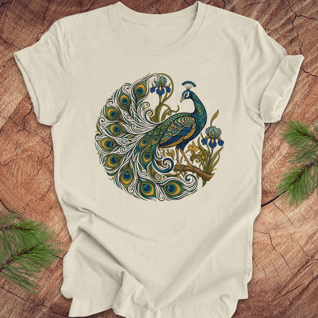 An beige short-sleeve t-shirt featuring a detailed graphic of a peacock with a fully fanned tail and blue flowers, laid flat on a rustic wood background with pine needles in the corners.