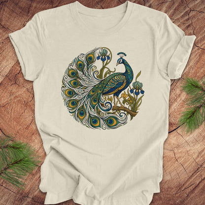 An beige short-sleeve t-shirt featuring a detailed graphic of a peacock with a fully fanned tail and blue flowers, laid flat on a rustic wood background with pine needles in the corners.