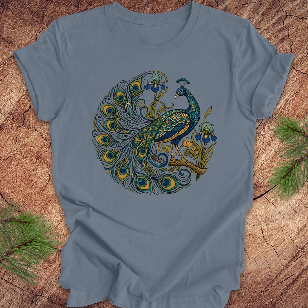 A light blue crewneck t-shirt featuring a detailed graphic of a peacock with a vibrant, multicolored tail and blue flowers, laid flat on a rustic wood background with pine needles.