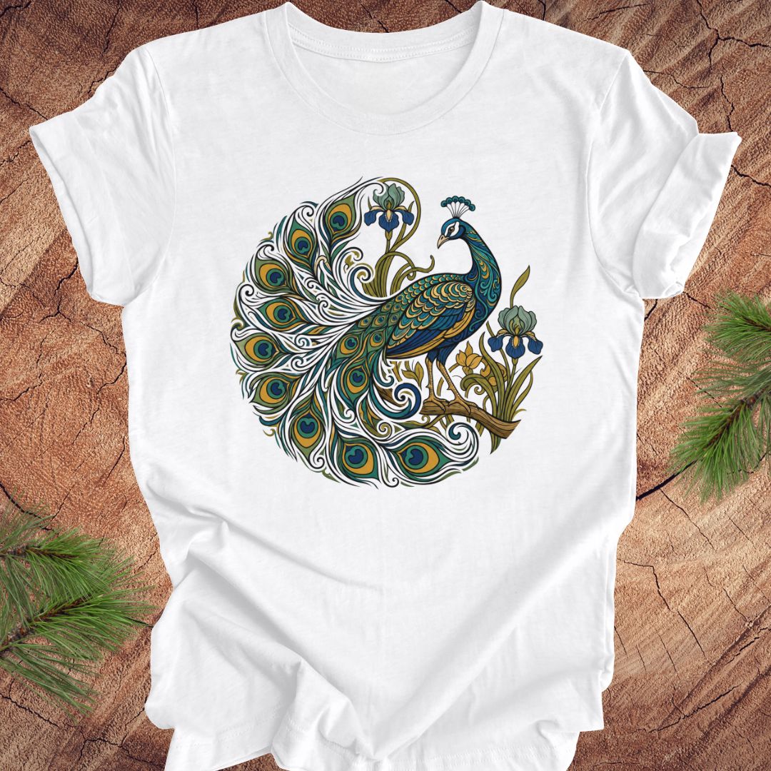 A white short-sleeve crewneck t-shirt featuring a detailed graphic of a peacock with its tail feathers spread in a circular pattern, styled flat on a rustic wood background with pine needles.
