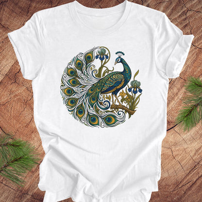 A white short-sleeve crewneck t-shirt featuring a detailed graphic of a peacock with its tail feathers spread in a circular pattern, styled flat on a rustic wood background with pine needles.