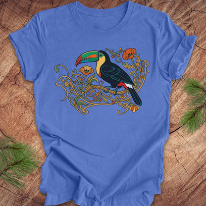 A blue short-sleeve crewneck t-shirt featuring a colorful graphic of a toucan perched on ornate gold vines, laid flat on a rustic wood background with pine needles in the corners.