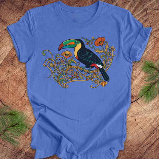 A blue short-sleeve crewneck t-shirt featuring a colorful graphic of a toucan perched on ornate gold vines, laid flat on a rustic wood background with pine needles in the corners.