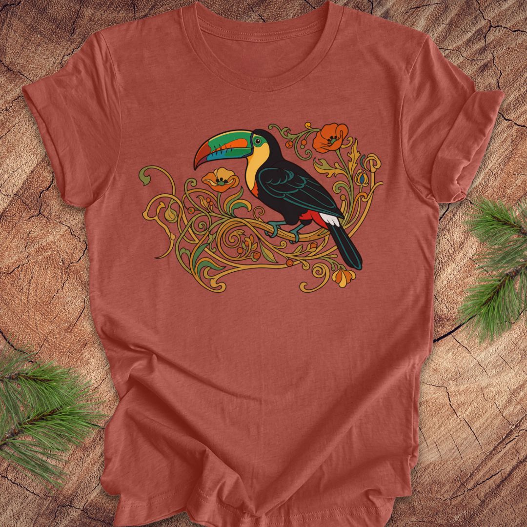 A terracotta-colored short-sleeve t-shirt featuring a colorful graphic of a toucan perched on ornate gold vines, laid flat on a rustic wood background with pine needles in the corners.