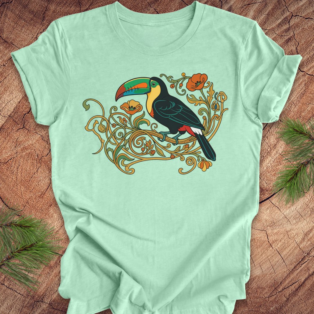 A mint green short-sleeve t-shirt with a colorful graphic of a toucan perched on ornate gold vines, laid flat on a rustic wood background with pine needles in the corners.