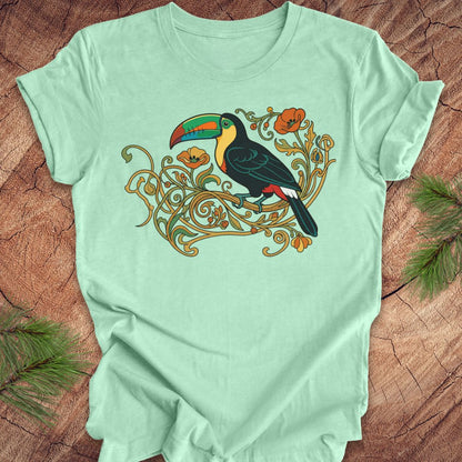 A mint green short-sleeve t-shirt with a colorful graphic of a toucan perched on ornate gold vines, laid flat on a rustic wood background with pine needles in the corners.