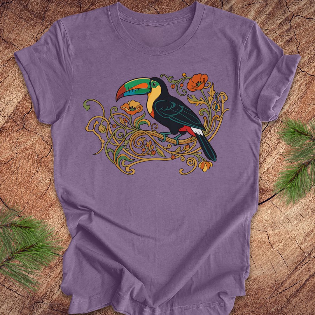 A purple short-sleeve t-shirt with a colorful graphic of a toucan perched on ornate gold vines, laid flat on a rustic wood surface with pine branches in the corners.