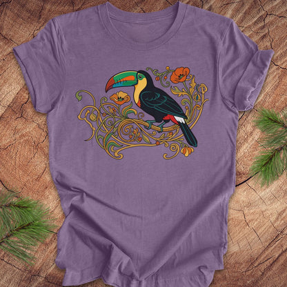 A purple short-sleeve t-shirt with a colorful graphic of a toucan perched on ornate gold vines, laid flat on a rustic wood surface with pine branches in the corners.