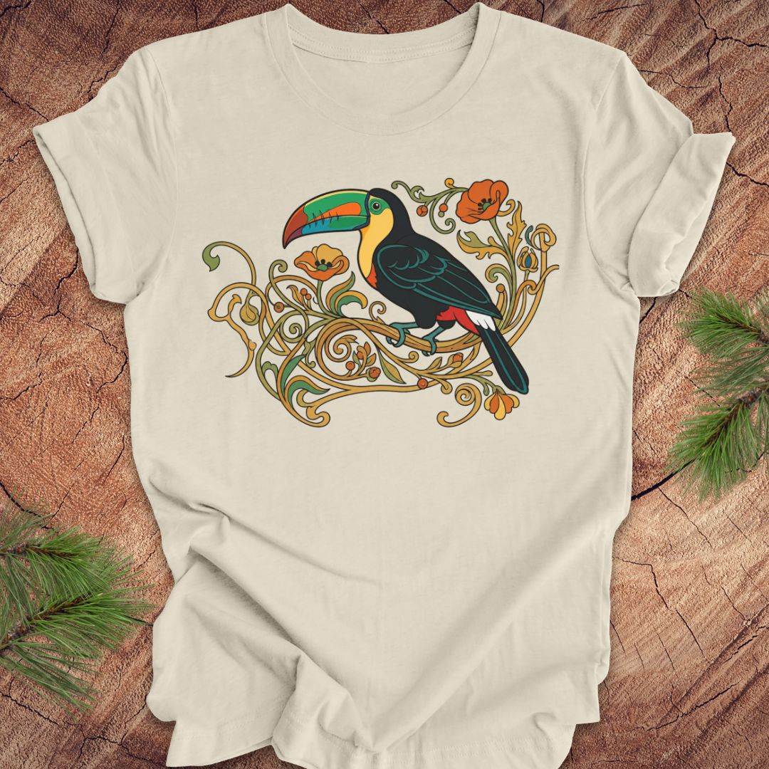 An beige short-sleeve t-shirt featuring a colorful graphic of a toucan perched on ornate gold vines with orange flowers, laid flat on a rustic wood background with pine needles in the corners.