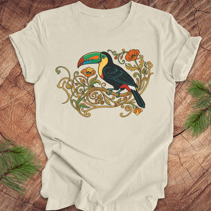 An beige short-sleeve t-shirt featuring a colorful graphic of a toucan perched on ornate gold vines with orange flowers, laid flat on a rustic wood background with pine needles in the corners.