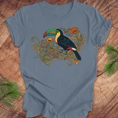 A slate blue crewneck t-shirt featuring a colorful graphic of a toucan perched on ornate gold vines, laid flat on a rustic wood background with pine needles in the corners.