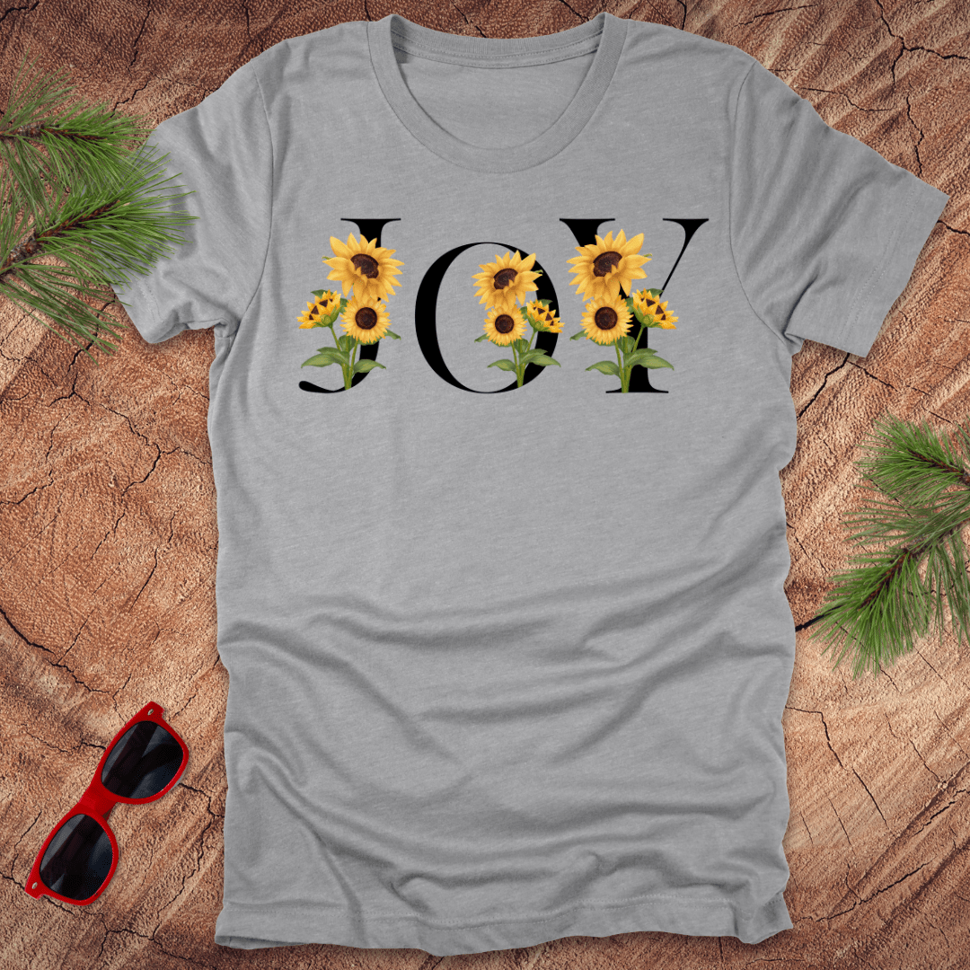 Gray t-shirt with 'JOY' and sunflower design on a wooden surface with sunglasses and greenery.