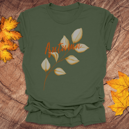 Green t-shirt with 'Autumn' design on a wooden surface with fall leaves.