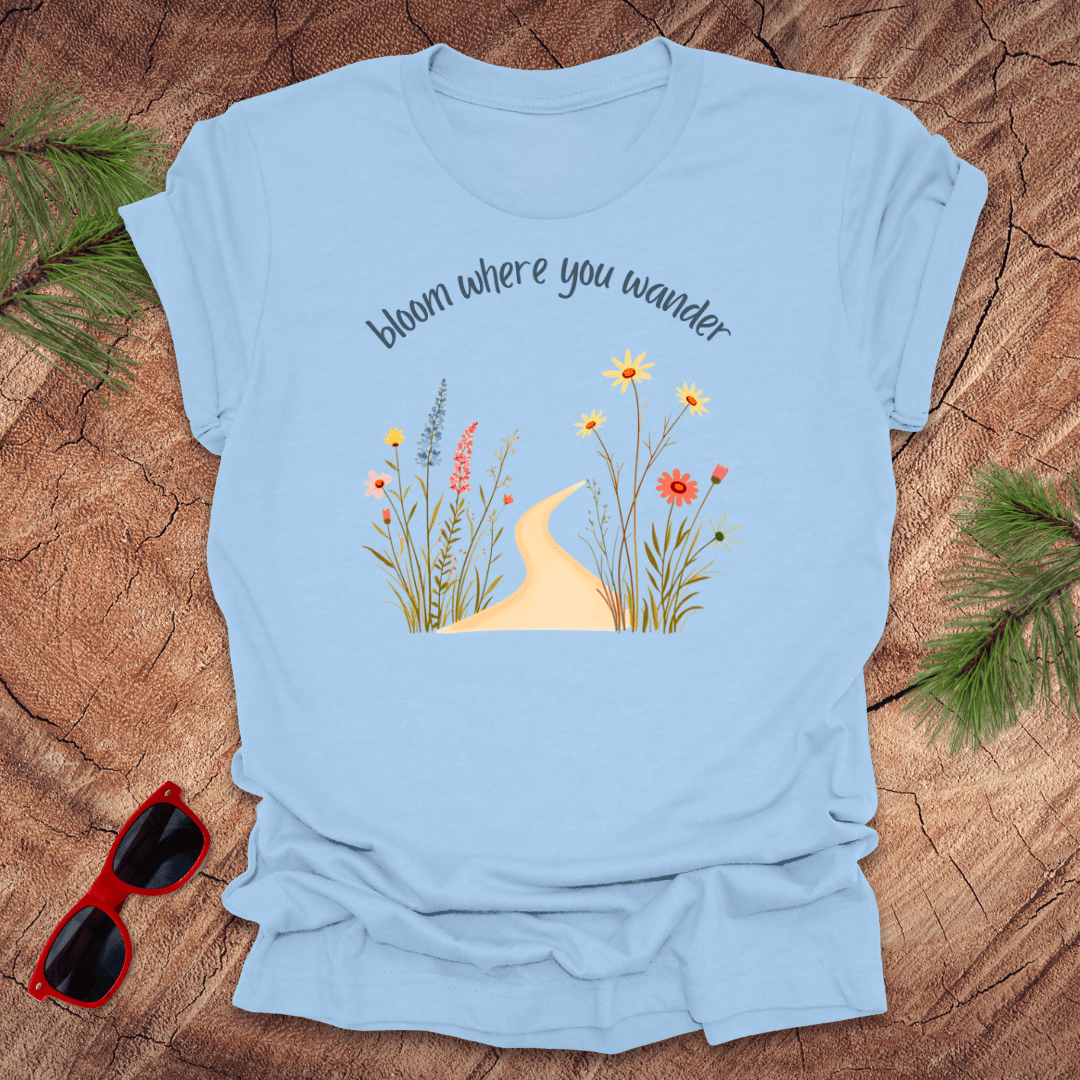 baby blue t-shirt with a graphic that includes a floral design and the text 'bloom where you wander', accompanied by a illustration of flowers on a wooden background with pine needles and red sunglasses to the side.