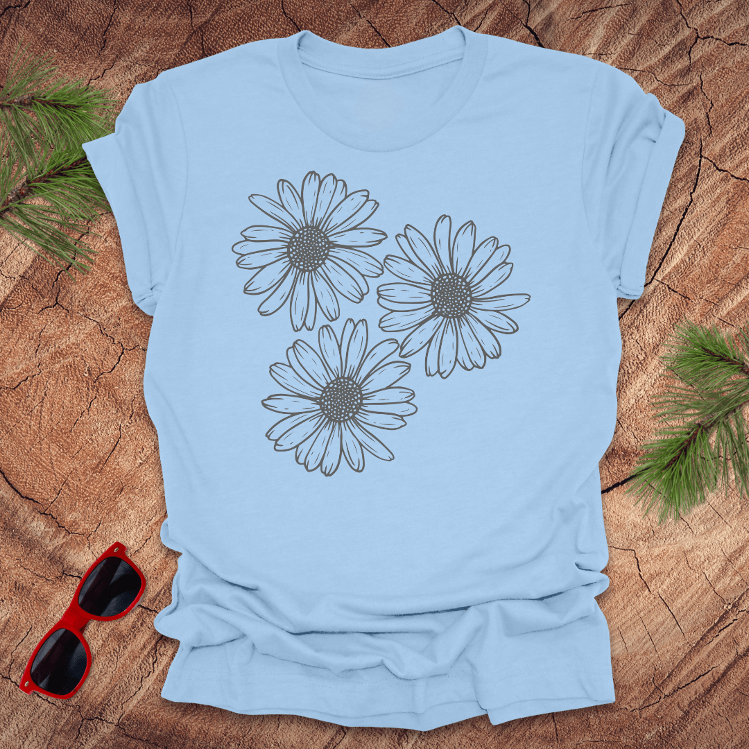 A baby blue t-shirt with 3 daisies prominently displayed. on a wooden surface with sunglasses and greenery.