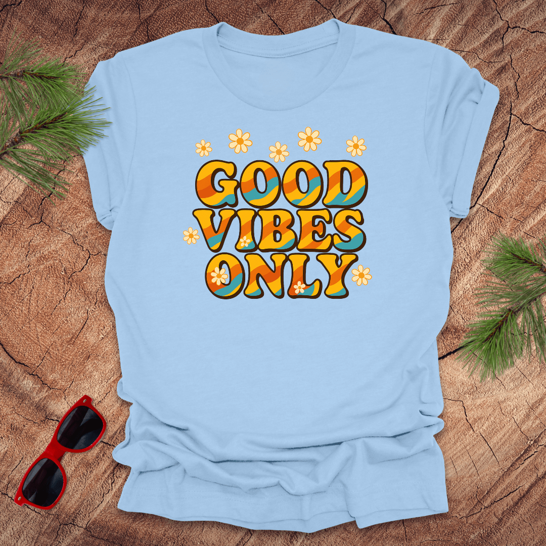Light blue t-shirt with 'Good Vibes Only' text on a wooden surface with sunglasses and greenery.