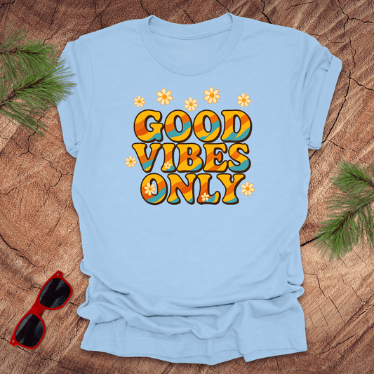 Light blue t-shirt with 'Good Vibes Only' text on a wooden surface with sunglasses and greenery.