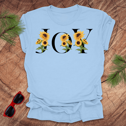 Light blue t-shirt with sunflower design and 'JOY' text on a wooden surface with sunglasses and greenery.
