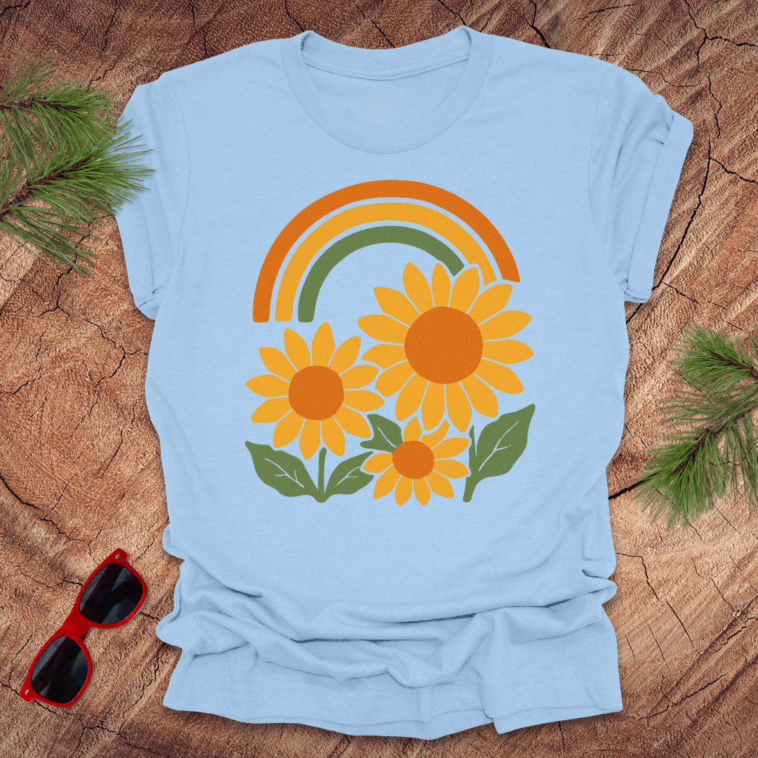 Light blue t-shirt with a rainbow and sunflower design on a wooden surface with sunglasses and greenery.
