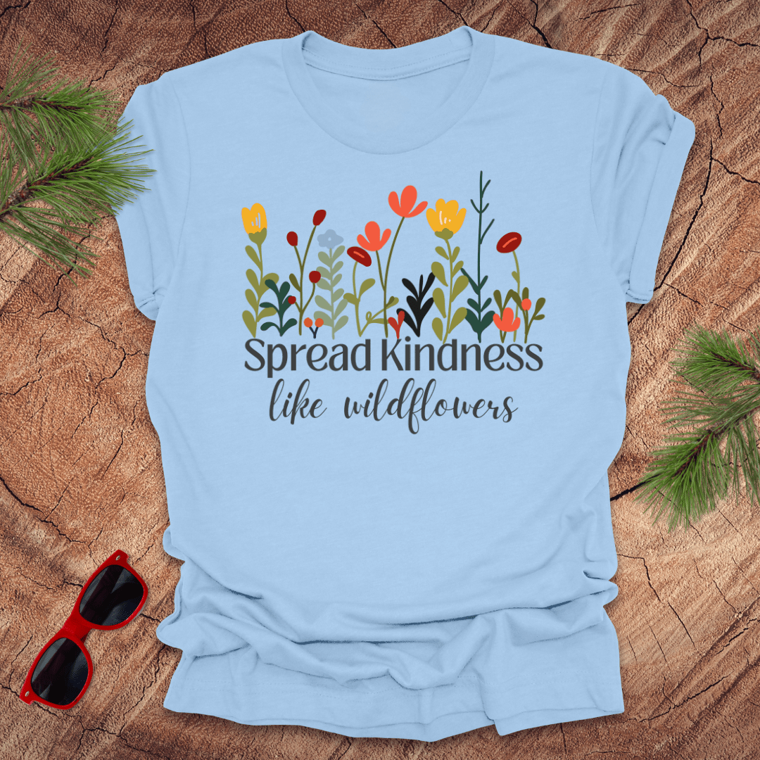Light blue t-shirt with floral design and text 'spread kindness like wildflowers' on a wooden surface with sunglasses and greenery.