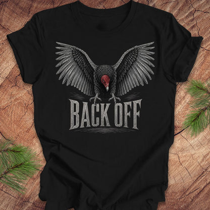 A black short-sleeve t-shirt laid flat on a rustic wood surface, featuring a large graphic of a spreading-wing vulture with a red head and the text 'BACK OFF' in distressed white lettering.