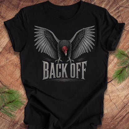 A black short-sleeve t-shirt with a graphic of a vulture with wings spread and the text 'BACK OFF' underneath, displayed on a rustic wood background with pine branches.