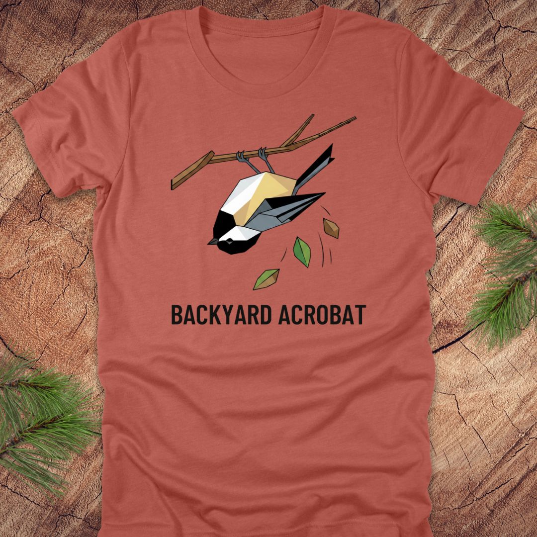 Clay colored t-shirt with a chickadee graphic and 'Backyard Acrobat' text on a wooden surface.