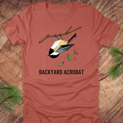 Clay colored t-shirt with a chickadee graphic and 'Backyard Acrobat' text on a wooden surface.