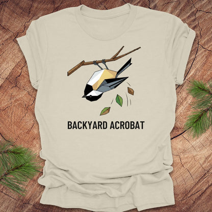 Beige t-shirt with a chickadee graphic and 'Backyard Acrobat' text on a wooden surface.