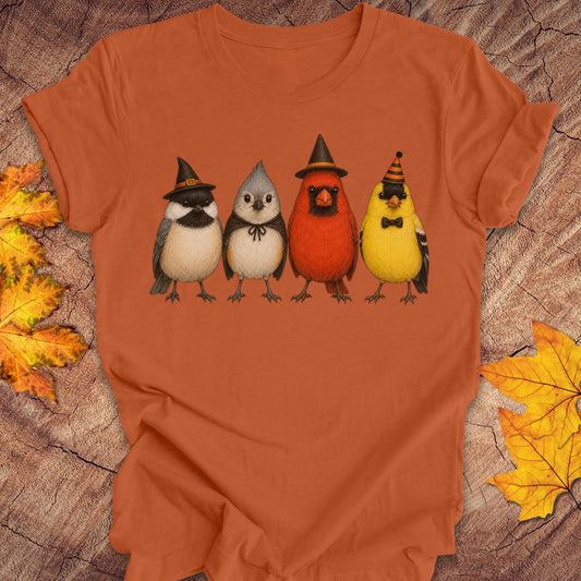 An orange short-sleeve crewneck t-shirt featuring a graphic of four cute birds in Halloween costumes (witch, owl, cardinal, and chick) on the chest, displayed flat on a rustic wood background with autumn leaves.