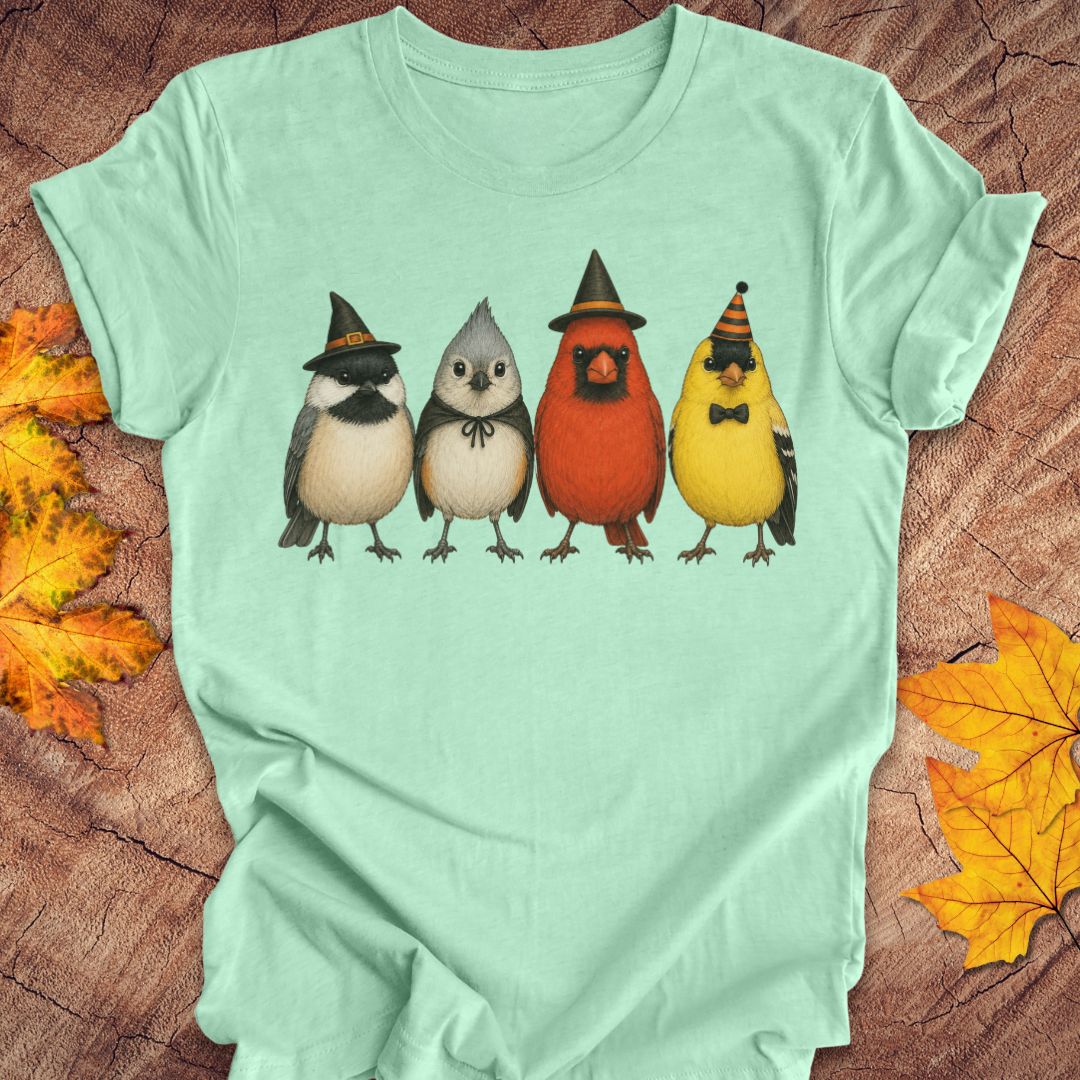 A mint green short-sleeve t-shirt laid flat on a rustic wood background with autumn leaves. The front of the shirt features a graphic of four cute birds wearing witch hats: a black and white bird, a gray bird, a red cardinal, and a yellow goldfinch.