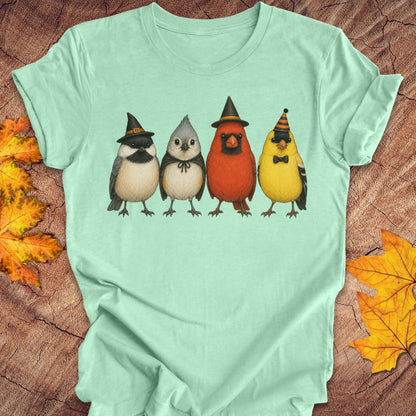 A mint green short-sleeve t-shirt laid flat on a rustic wood background with autumn leaves. The front of the shirt features a graphic of four cute birds wearing witch hats: a black and white bird, a gray bird, a red cardinal, and a yellow goldfinch.
