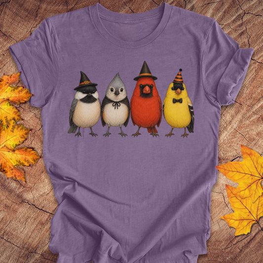 A purple short-sleeve t-shirt laid flat on a rustic wood background with autumn leaves. The shirt features a graphic of four cute birds wearing witch hats: a bluebird, a gray owl, a red cardinal, and a yellow chick.