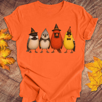 An orange short-sleeve t-shirt laid flat on a rustic wood surface, featuring a graphic of four cute birds wearing witch hats and sunglasses. The background is decorated with scattered autumn leaves.