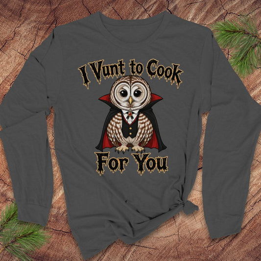 A dark gray long-sleeve t-shirt with a graphic of a vampire owl and the text 'I Vunt to Cook For You' laid flat on a rustic wood background with pine branches.